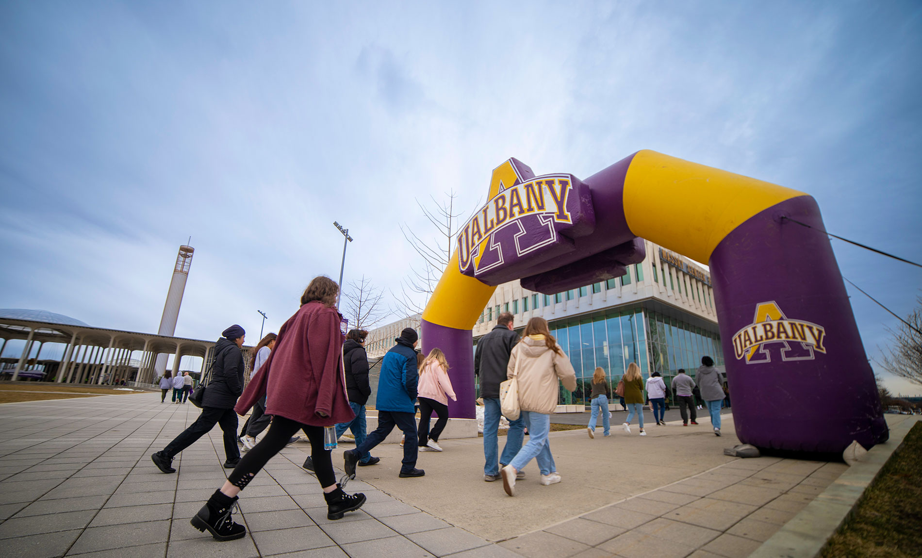 A group of people walking under an inflatable purple and gold arch with the UAlbany logo on it.