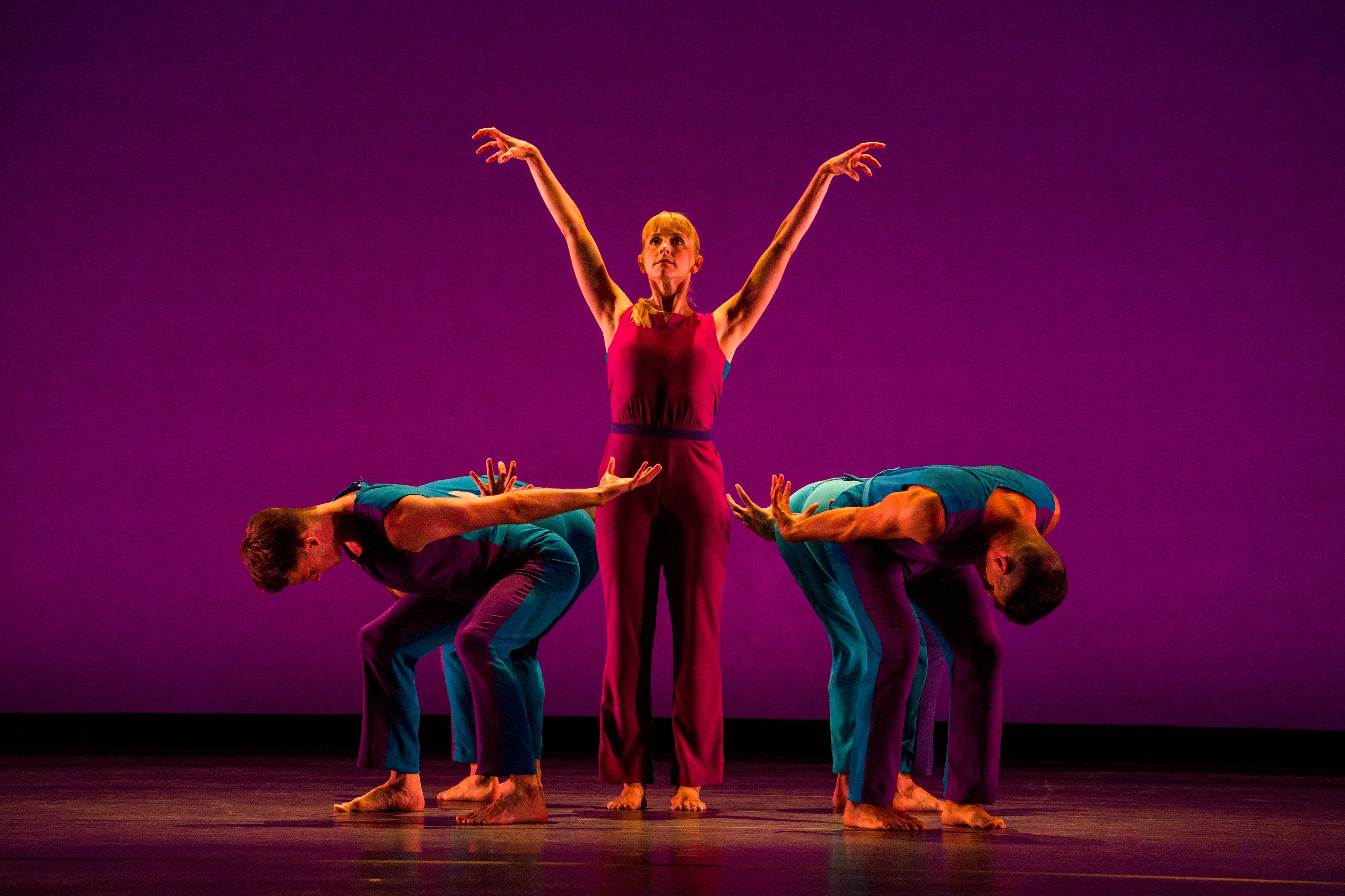  a dancers stands center with arms raised overhead while other dancers bend forward and away
