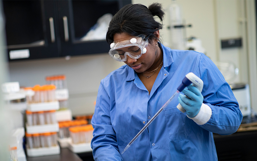 A woman in goggles and a blue lab coat uses lab equipment while conducting research