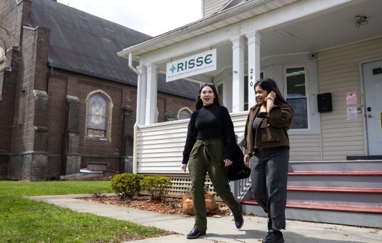 Two women smile as they exit a building with a sign reading: “RISSE Refugee & Immigrant Support Services of Emmaus” hanging from the porch roof. A brick church is visible in the background.