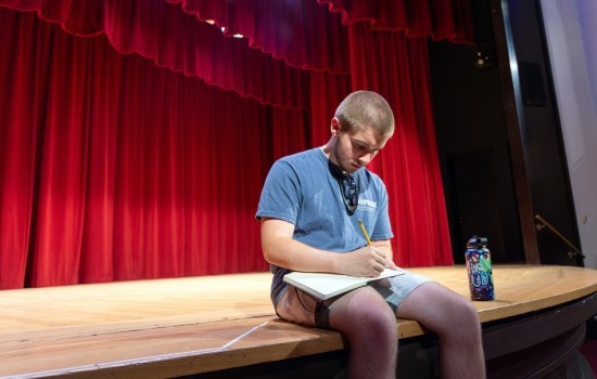 A teen boy sits on a theatre stage in front of a red curtain and writes in a notebook.