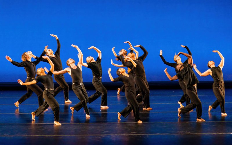 many dancers costumed in black lean back looking upward with arms extended high