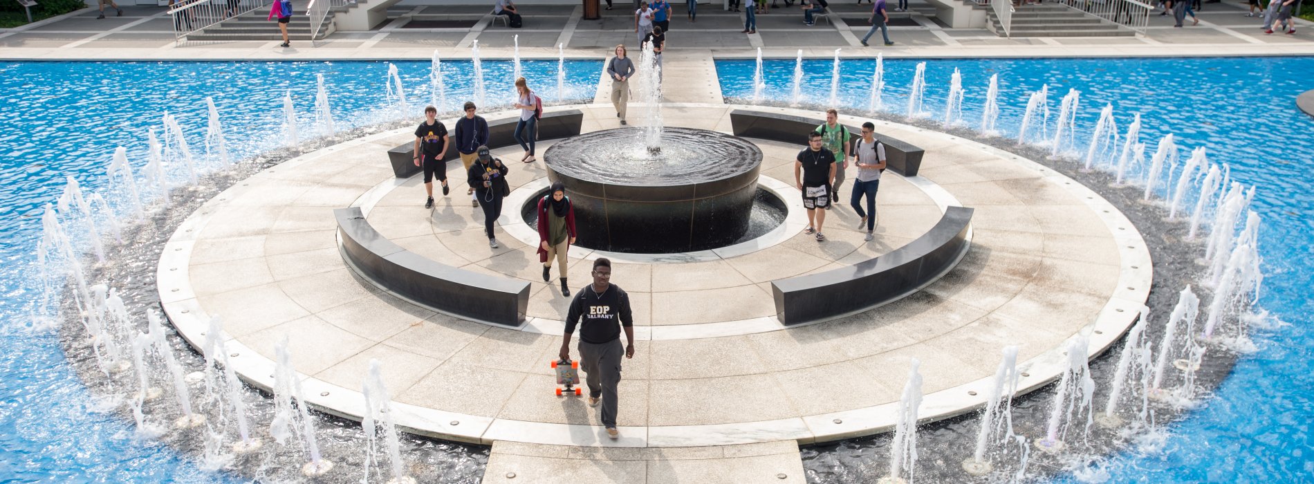 Students walk through UAlbany's academic podium, which includes staircases, overhangs, benches and a fountain.