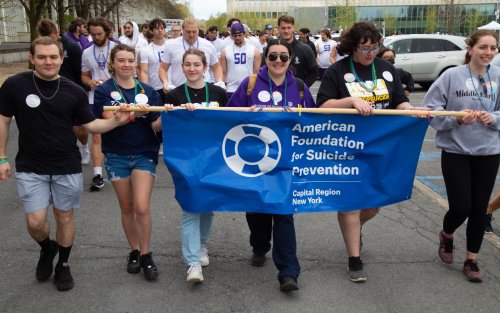 Photo depicts students leading UAlbany's 2023 "Out of the Darkness" walk. In front are six students holding a blue banner reading "American Foundation for Suicide Prevention, Capital Region New York". They are followed by a crowd of walkers. Brianna Lennon is among the banner holders, wearing a purple hoodie and dark aviator sunglasses.