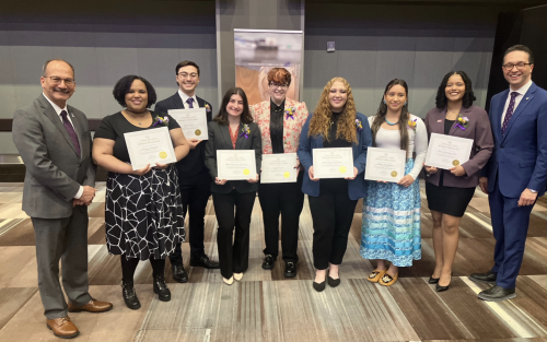 Seven students in professional attire stand holding award certificates and smile for a picture with UAlbany President Havidán Rodríguez and Vice President for Student Affairs Michael Christakis in a conference room.