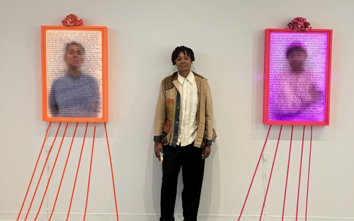 A person with short black braids, a tan jacket, white button down and black slacks stands against a white wall in between two lightbox portrait outined in red and orange and covered with text.