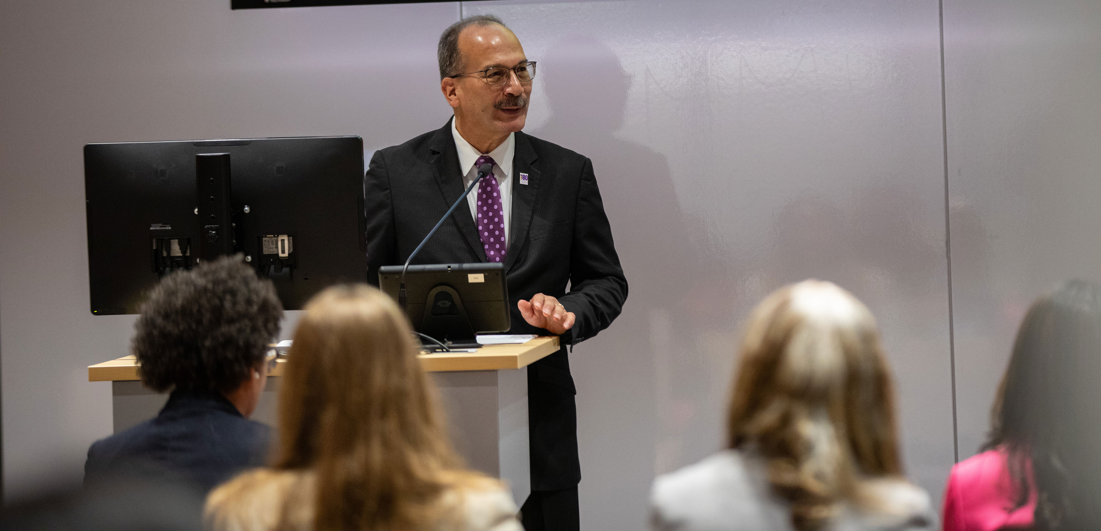 UAlbany President Havidán Rodríguez speaks at a podium with a computer in front of a room of people.