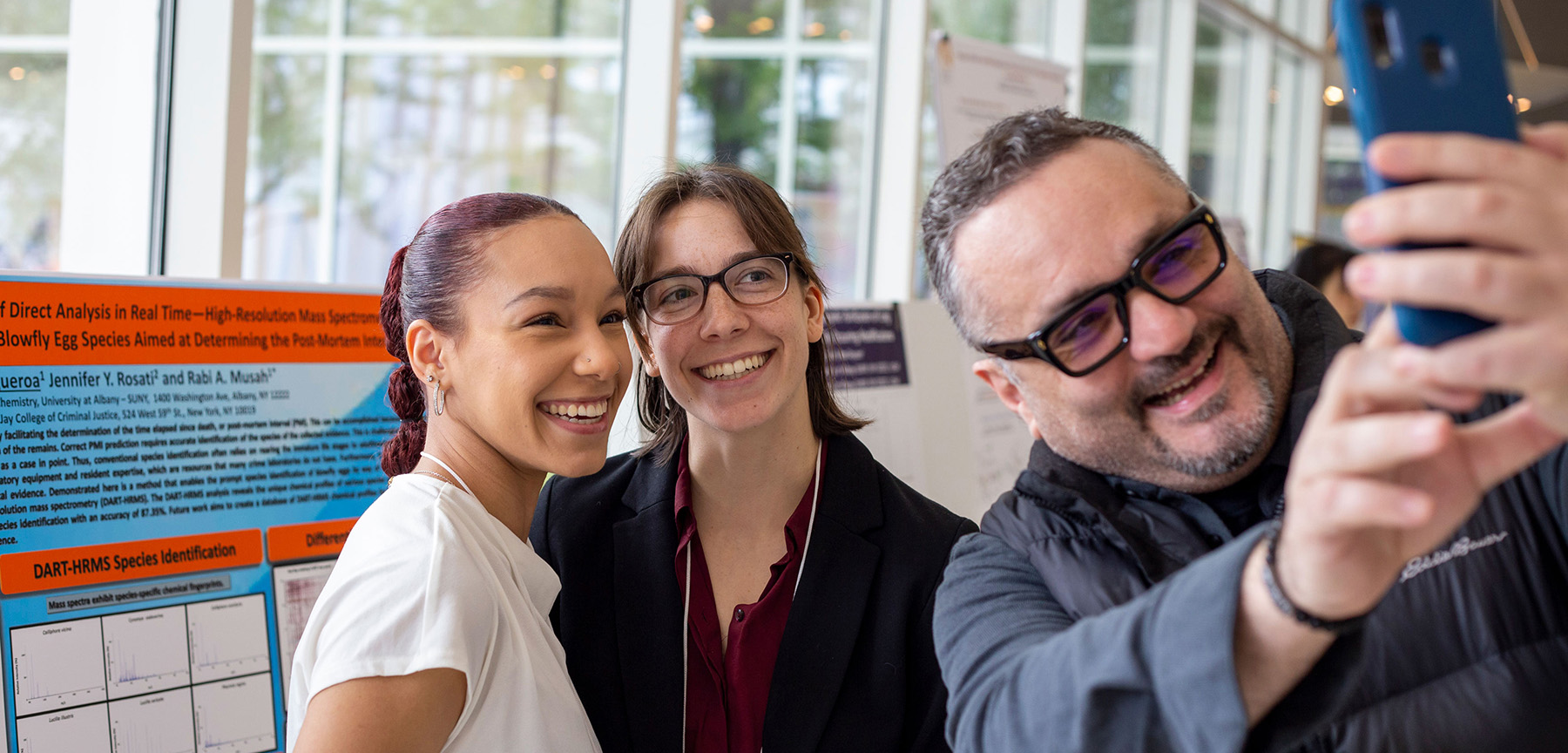 A faculty member smiles as he uses his cell phone to take a selfie with two smiling students posed in front of a research poster.