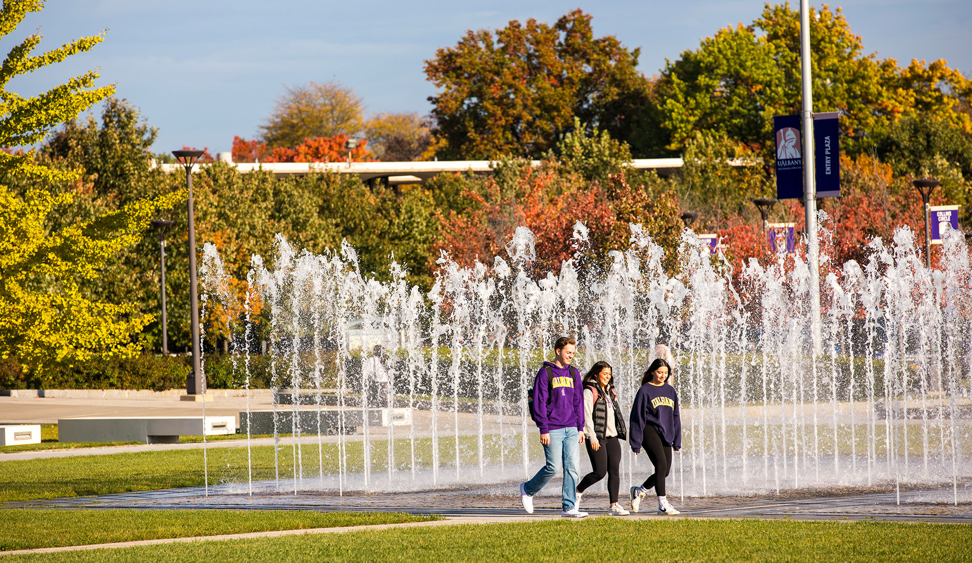 Three students walking by the entrance fountain together in the fall.