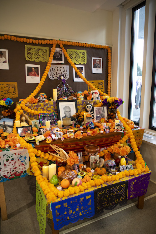 A colorful ofrenda is decorated with orange paper marigolds, candles, painted skulls and skeletons, food, and pictures of loved ones inside the Arts & Sciences building.