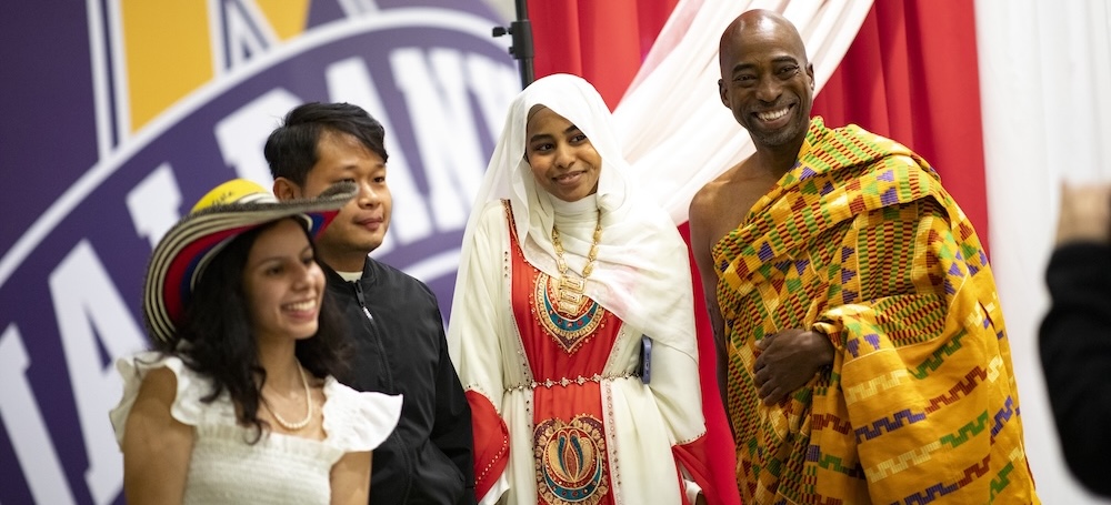 Four people from varied cultures stand before a large UAlbany banner.