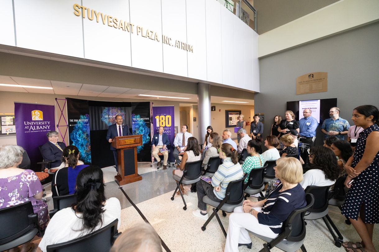 Image shows a bright sunlit room full of people listening to a man wearing a navy suit and purple tie. Many are smiling and applauding. 