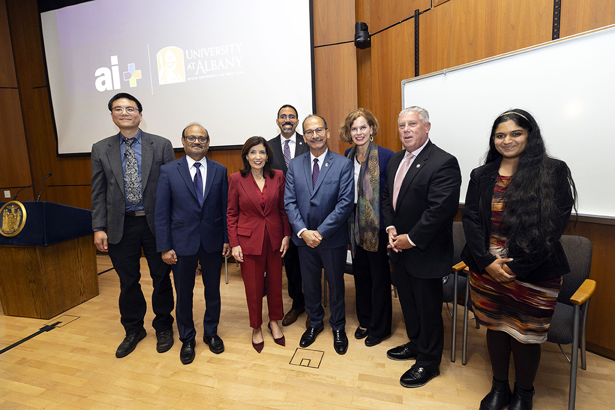 Group of seven individuals standing in front of University at Albany and AI logos, smiling in a conference room setting.