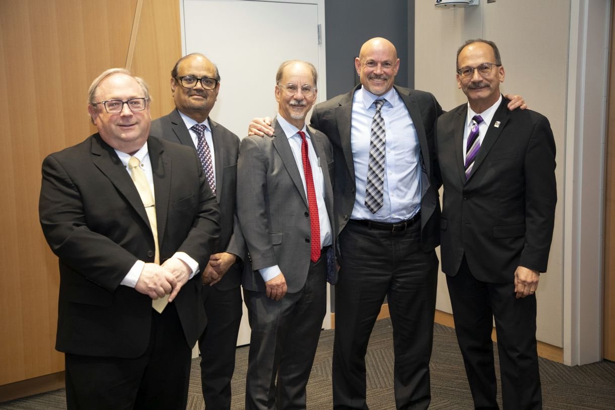 Five men wearing suits, all with ties and four with glasses, smile together for a casual group portrait.