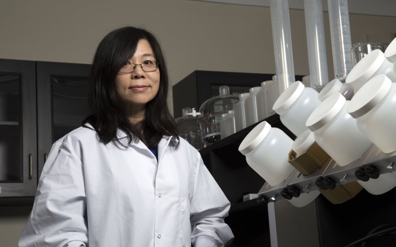 A portrait of a woman in a white lab coat standing a lab surrounded by various metal and plastic containers.