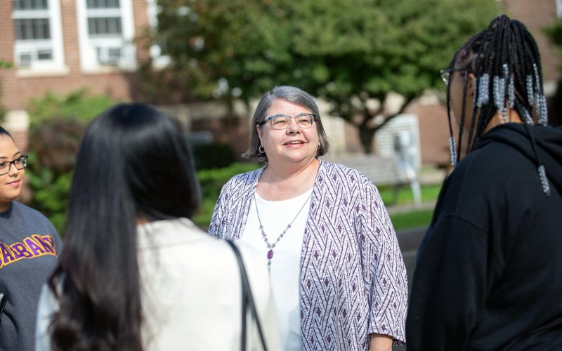 Four people stand together outdoors. A woman in the center smiles at three female students who are facing her. The woman has short gray hair and is wearing a purple and white patterned jacket and glasses. A tree and red brick building are in the background.