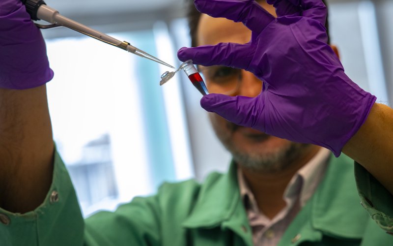 A man wearing a green lab coat and purple gloves holds a pipette and a small vial of solution. The top of the solution is red and the bottom of the solution is purple.