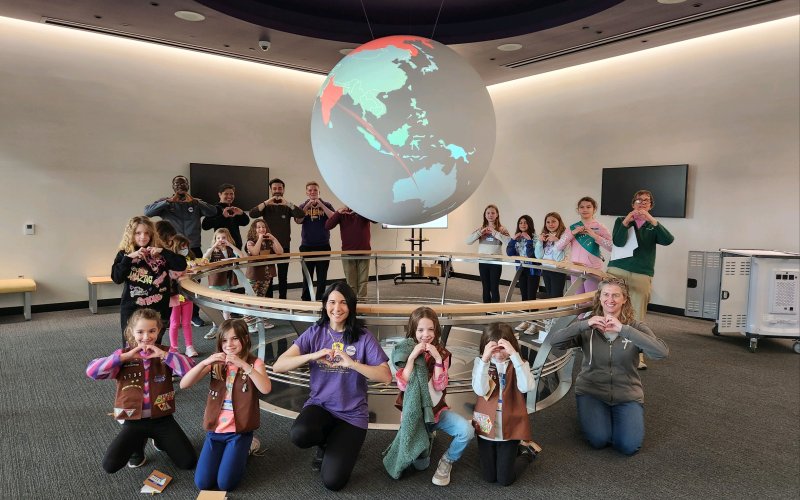 A group of people young and old standing and kneeling around an illuminate globe making heart symbols with their hands.