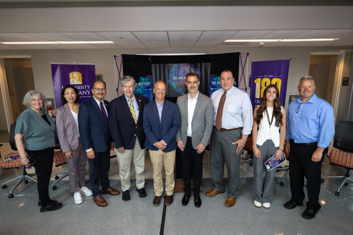 Nine people (three women and six men) stand together in a row, smiling for a group photo. They are standing in a row in front of three banners bearing the UAlbany and RNA Institute logos set up for a press conference in the Life Science Research Building atrium.