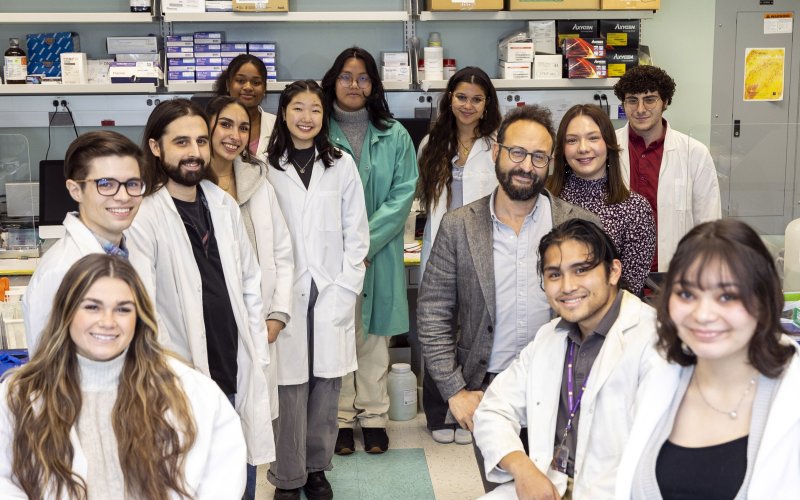 Image shows 13 smiling people, including a mix of genders, in a brightly lit science lab. Most are undergraduate and graduate-level students wearing white lab coats. Shelves on the back wall hold boxes of lab supplies.