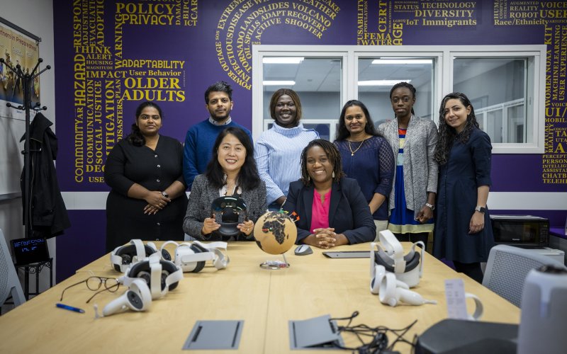 CEHC's Extreme Events, Social Equity and Technology research team stand in front of a purple and gold branded wall inside their lab space.