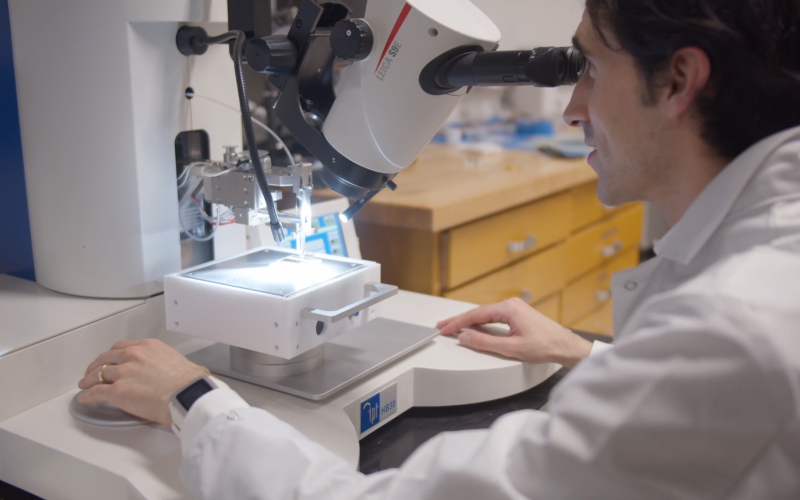 A dark-haired man in a white lab coat is seated as he looks into the eye piece of some lab equipment.