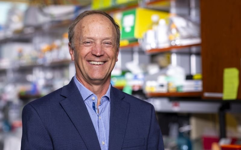 Portrait of Andy Berglund: A smiling man wearing a blue blazer stands in a laboratory. The background, which is blurred, includes shelves full of lab supplies. 