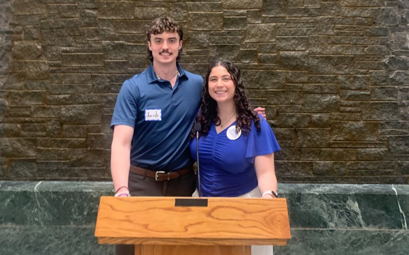 Students Jacob Schoff and Alexis Candido pose for a photo behind a wooden podium in the NYS Capitol building. Both are smiling and wearing blue short sleeved shirts. The wall behind them is made of brown stone and green marble.