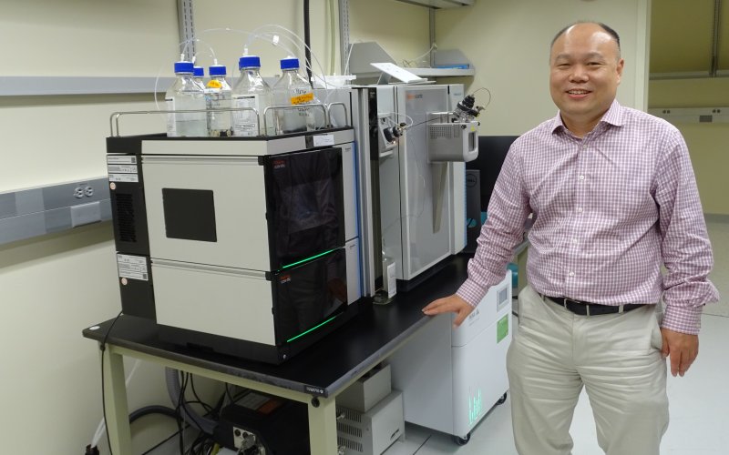 A man wearing khaki pants and a plaid button-down shirt smiles in a brightly lit lab. He is standing next to a DNA/RNA sequencing machine placed on top of a lab bench. The machine consists of two large metal cube-shaped containers. Five bottles of clear liquid stand on top of one; the other has a digital panel facing the front. 