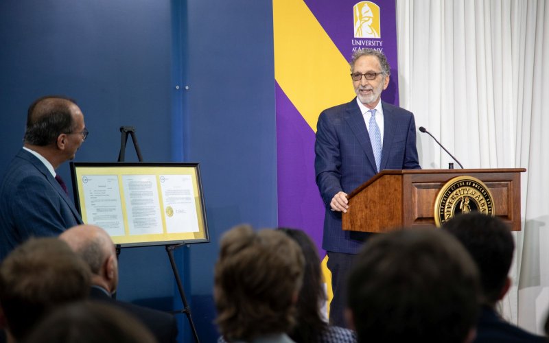 A man in a blue suit stands at a lectern speaking before a crowd while another man looks on from the side.