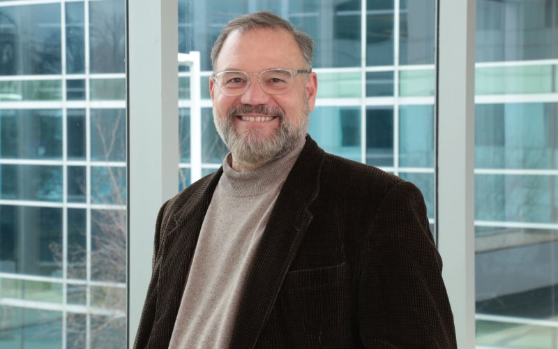 Portrait of Scott Tenenbaum: A man wearing clear-rimmed glasses, a dark brown blazer and tan turtleneck sweater stands, smiling, in front of a wall of glass windows.