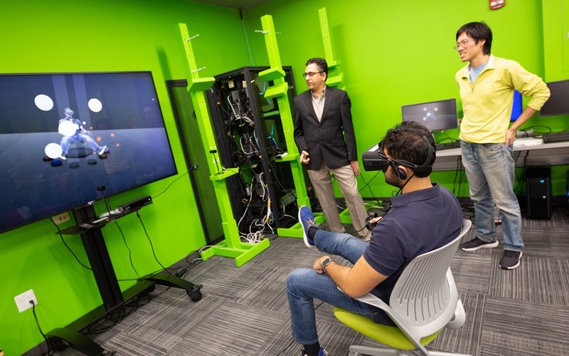 A man wearing a VR headset is seated in a chair facing a large video monitor while two other men stand nearby watching.