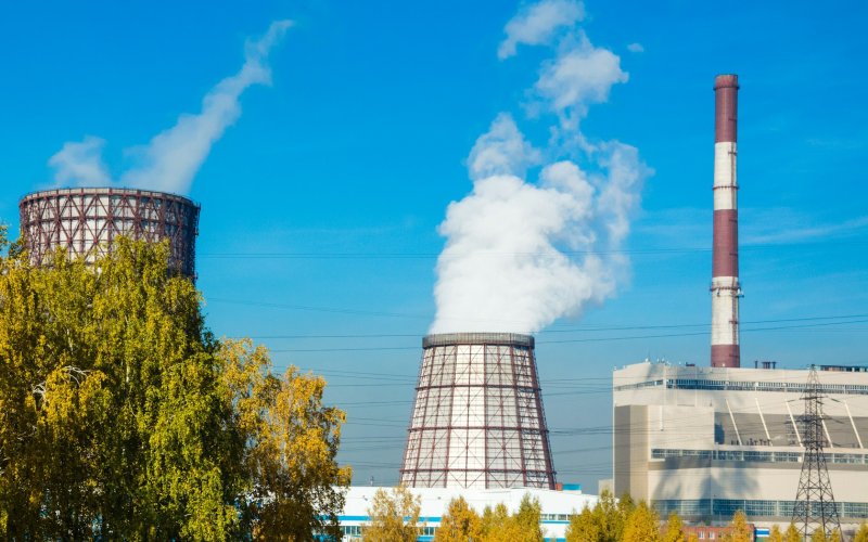 Landscape image featuring two large, conical industrial cooling towers near a gray building with a tall red and white smokestack. Trees are in the foreground and plumes of mist from the two towers drift skyward.