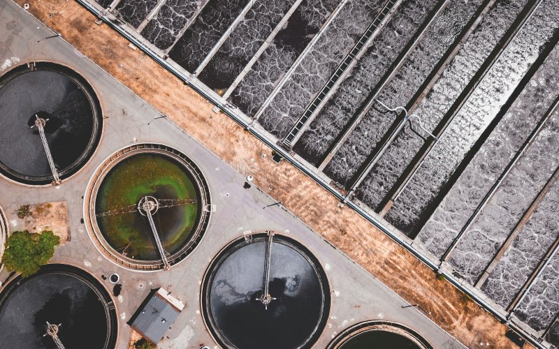 Aerial image of a wastewater treatment facility. The top right half of the image shows water being treated in a rectangular holding pool divided into parallel segments. On the bottom left, water is held in circular tanks.