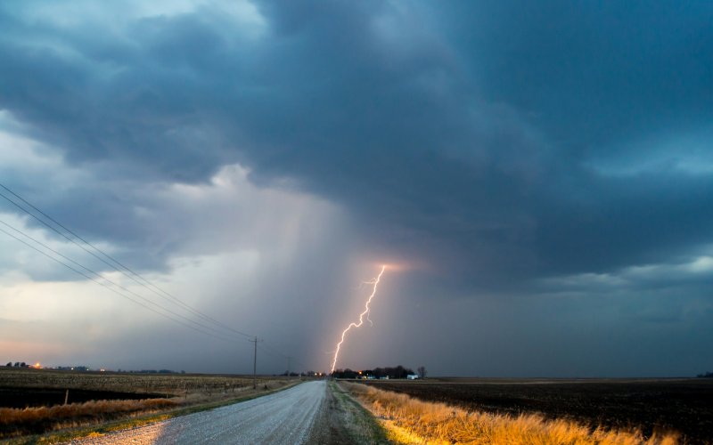 Image shows a rural landscape beneath a dark, clouded sky. A lightning strike touches down near an isolated settlement in the distance. Powerlines run along a gravel road leading to the settlement.