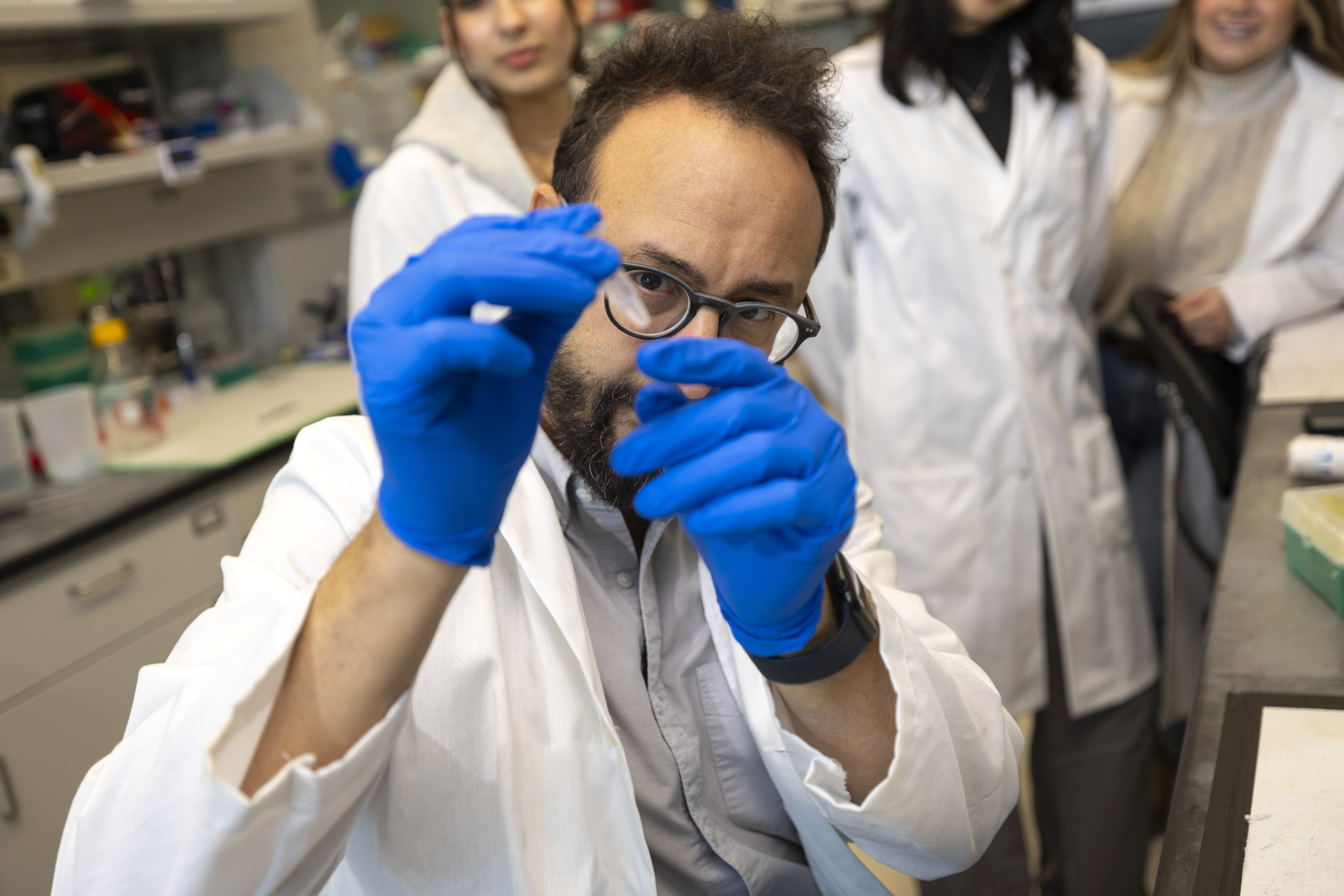 A man with glasses and blue gloves hold up a small plastic test tube filled with clear liquid.