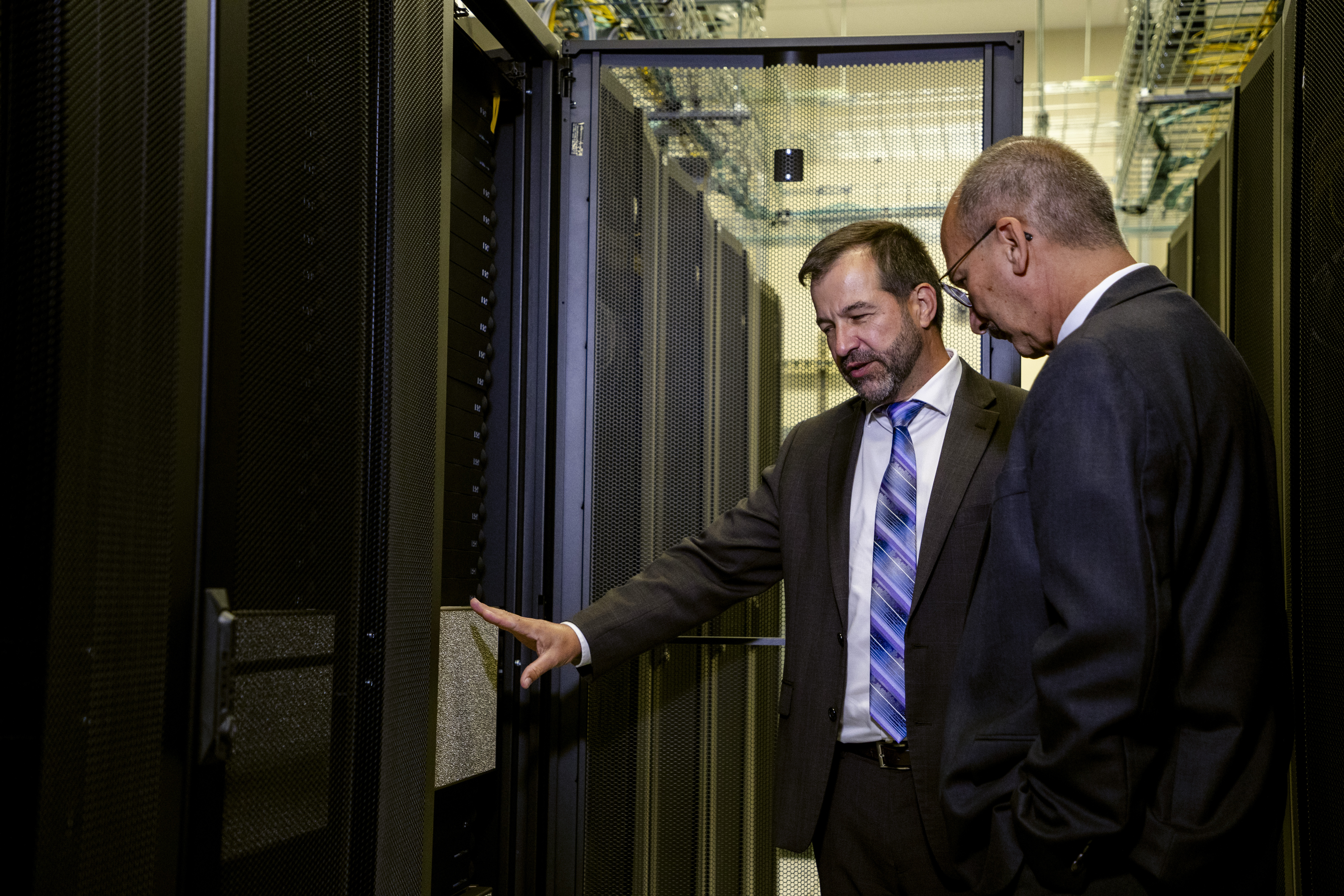 A man in a dark suit and purple tie gestures toward a black computer server rack.