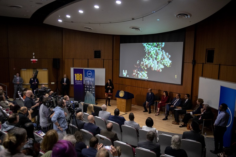 A crowded seared in an auditorium watches as a women explains the images of colorful molecules projected on a large screen.