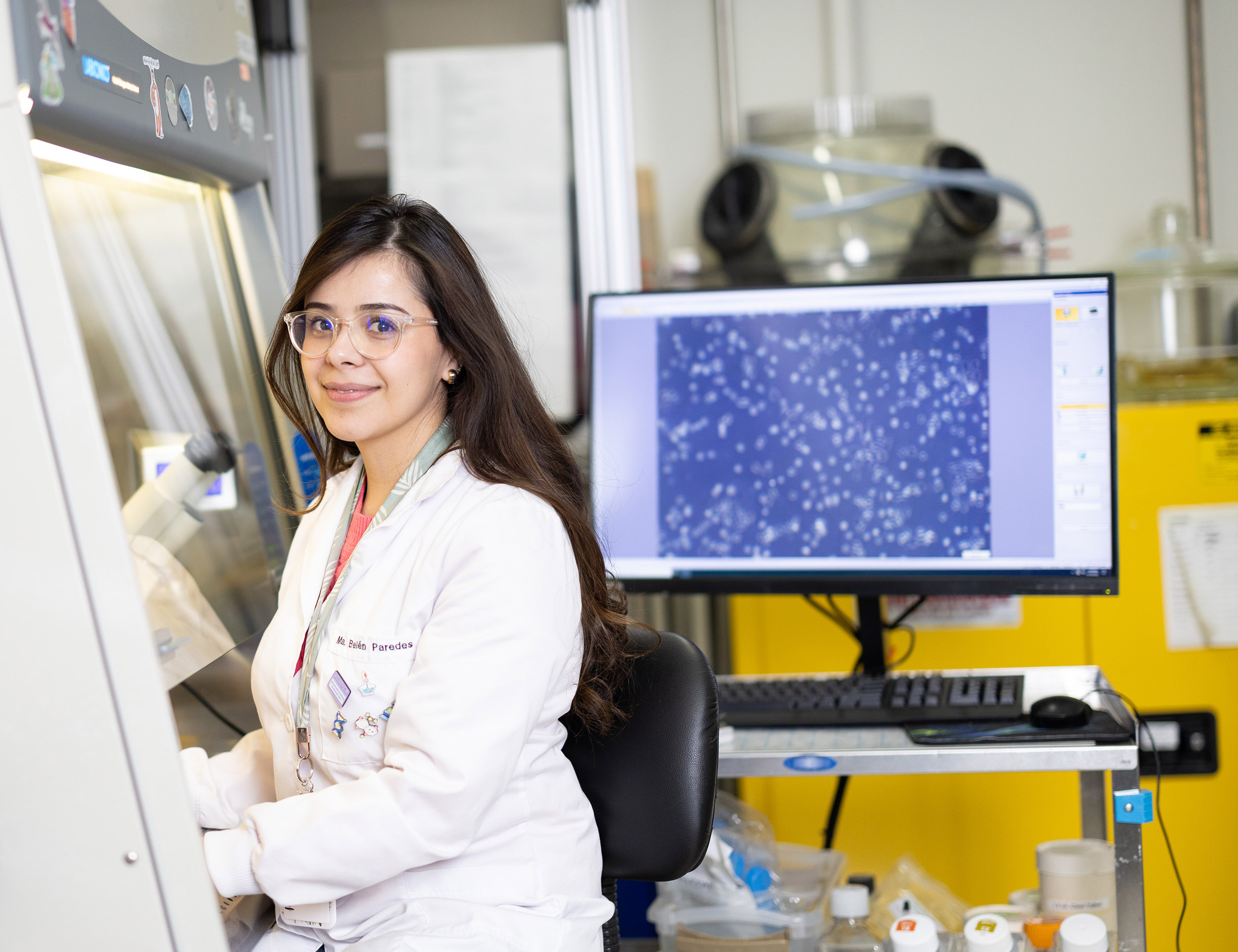 A young woman with dark hair and glasses wearing a white lab coat sits at a research station under a hood. Behind her is a monitor with a blue screen and tiny heart cells on display.