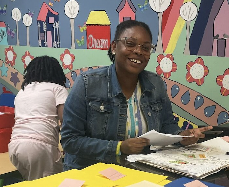 A woman in a jean jacket holds papers and smiles as she is seated at a table topped with colorful folders in a classroom with kids.