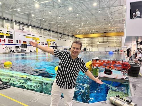 A man in a pinstripe shirt poses with his arms outstretched in front of a large pool at the Neutral Buoyancy Laboratory.