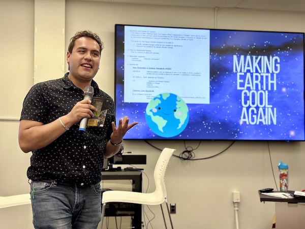 A young man in a dark button-down shirt speaks into a microphone in a classroom in front of a screen that reads "Making Earth Cool Again"