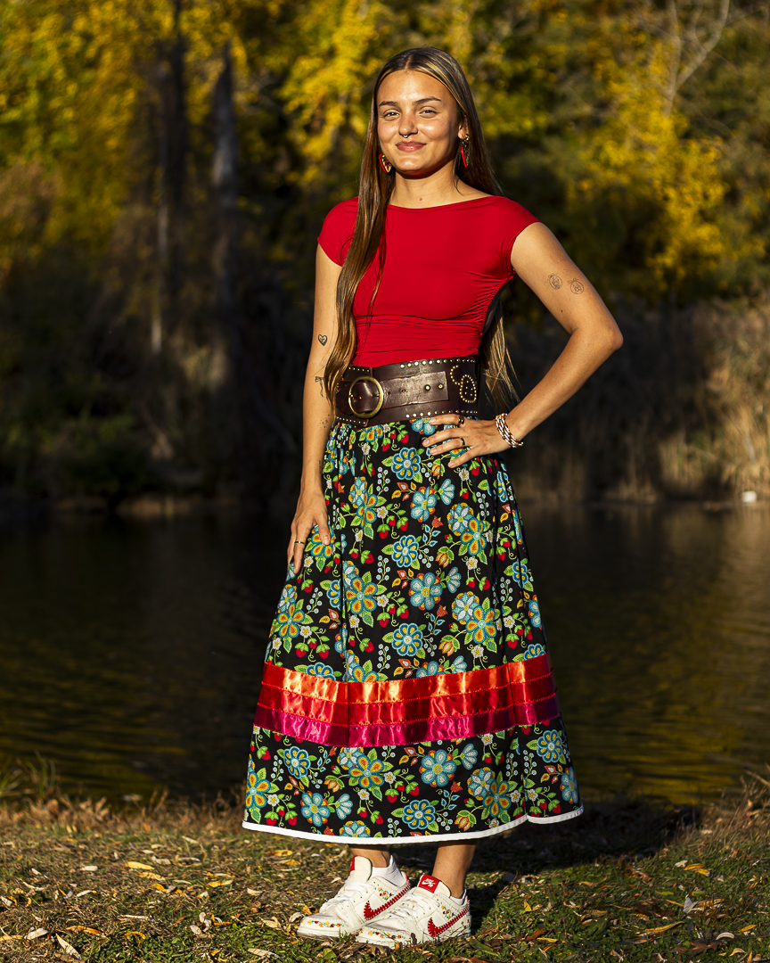 A young woman with long brown hair, a red shirt and colorful long skirt poses for a portrait outdoors on a sunny fall day.