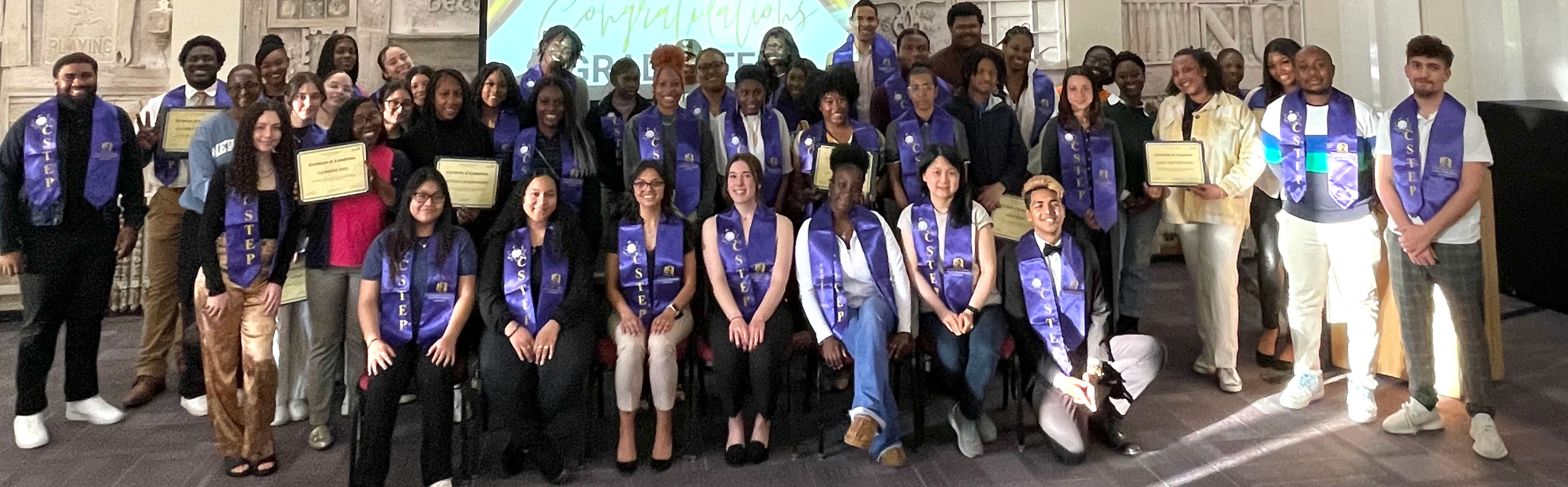 Dozens of students pose for a group photo while wearing their purple CSTEP graduation stoles. Several students hold up certificates proudly.