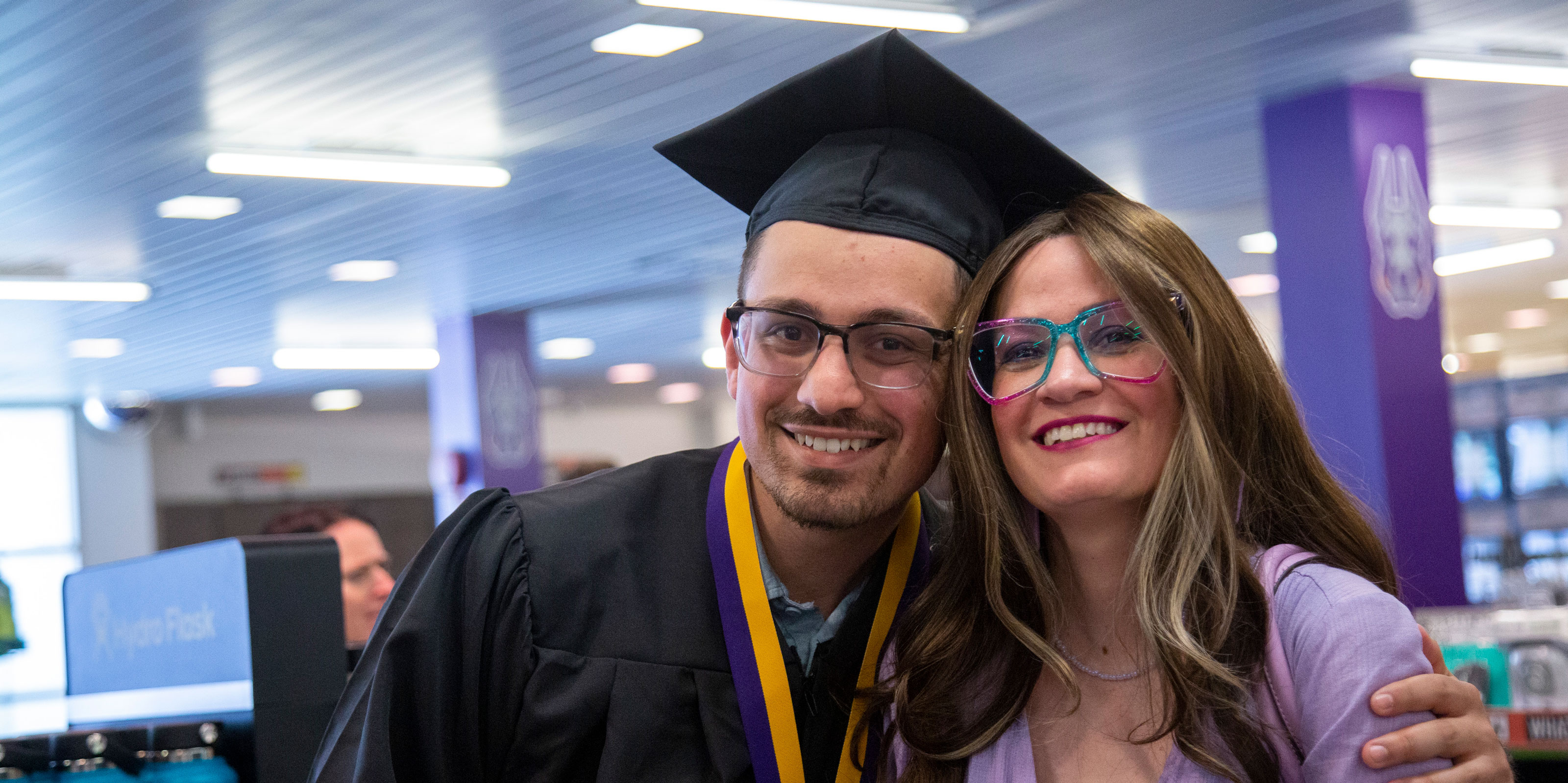 A UAlbany graduate wearing Commencement cap and gown poses with a loved one inside the campus bookstore.