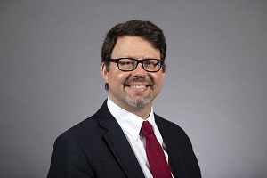 A portrait of a man with dark hair and glasses wearing a dark suit, white shirt and red tie smiling toward the camera.