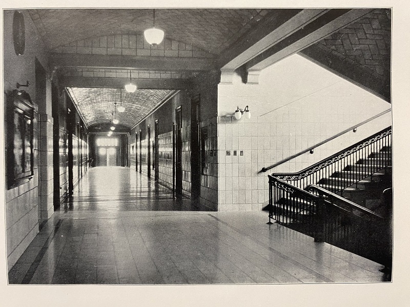 A black and white photo of a long, back-lit hallway with intricate tile ceilings.