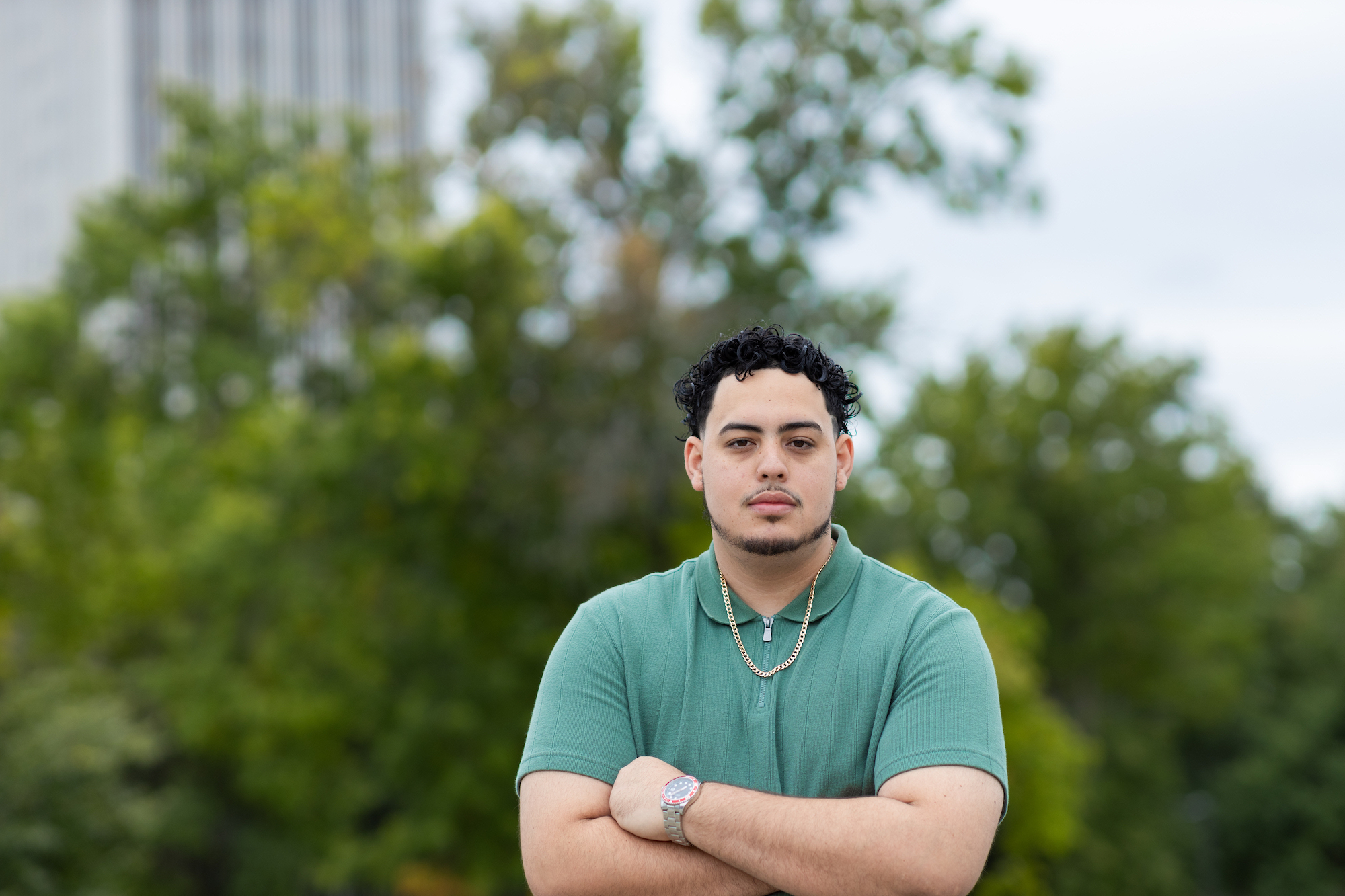 A young man with short curly black hair and a mustache wearing a green polo shirt and chain poses outdoors with his arms crossed.