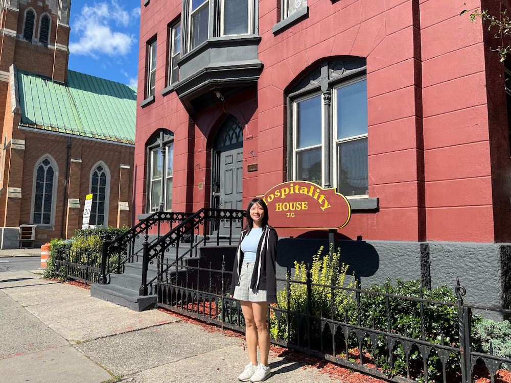 A young woman stands outside of a red building on a sunny day in front of a sign that says "Hospitality House"