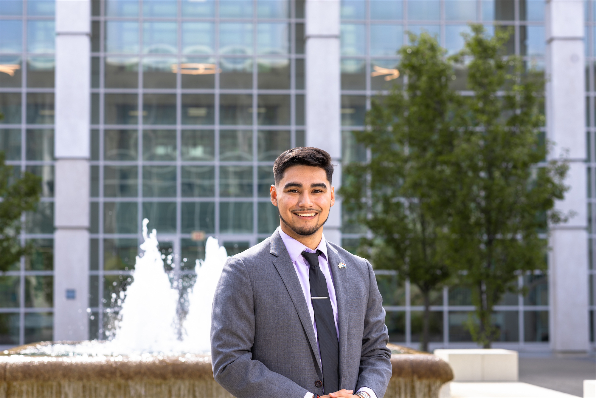 A young man with short black hair and a goatee wearing a gray suit and black tie smiles and poses for a portrait in front of a fountain. 